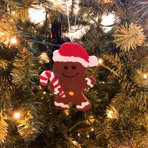 Puede incluir: Un adorno de hombre de jengibre marr&oacute;n con un gorro de Pap&aacute; Noel rojo y blanco, sosteniendo un bast&oacute;n de caramelo a rayas rojas y blancas. La figura presenta detalles de glaseado blanco en los brazos y piernas, con un bot&oacute;n verde y uno amarillo.