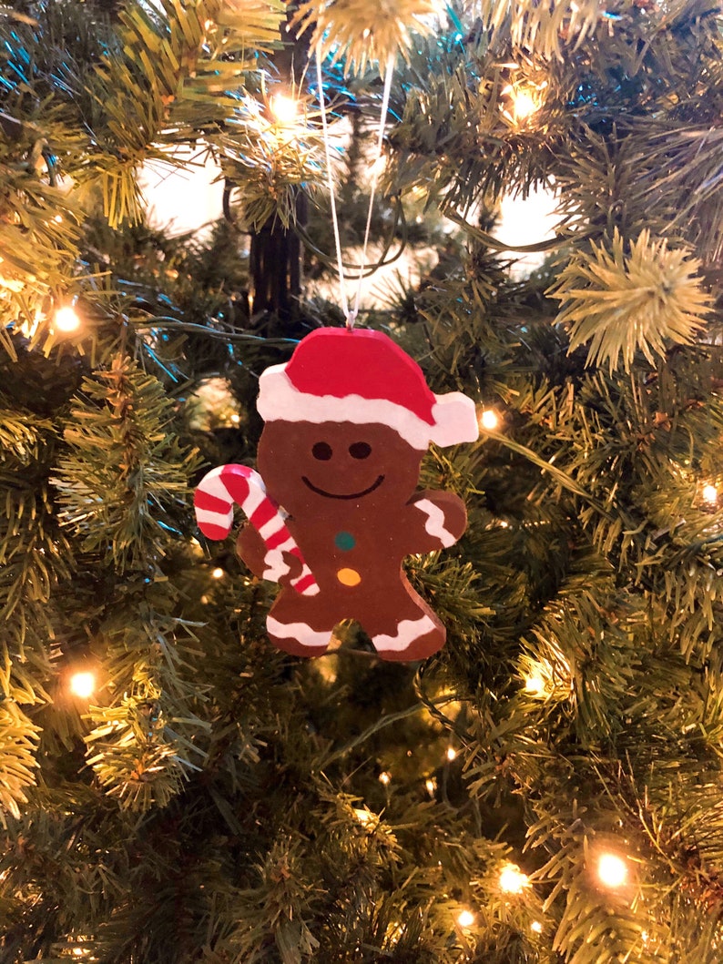 Puede incluir: Un adorno de hombre de jengibre marr&oacute;n con un gorro de Pap&aacute; Noel rojo y blanco, sosteniendo un bast&oacute;n de caramelo a rayas rojas y blancas. La figura presenta detalles de glaseado blanco en los brazos y piernas, con un bot&oacute;n verde y uno amarillo.