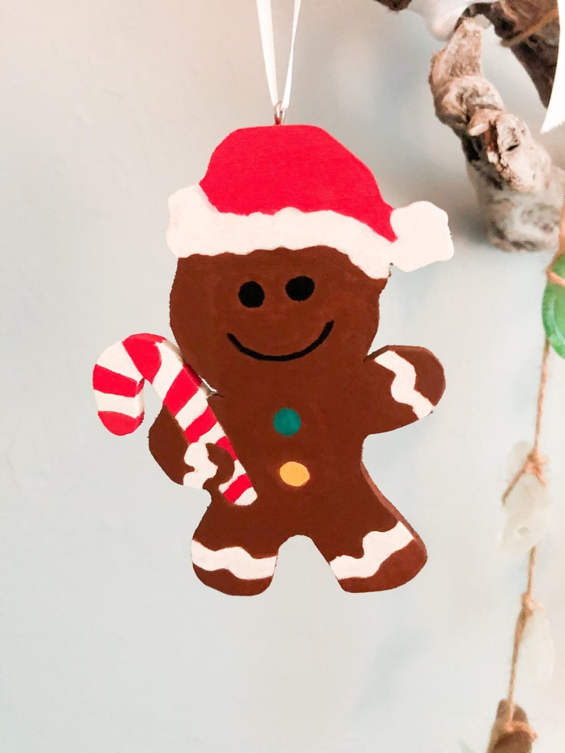 Puede incluir: Un adorno de hombre de jengibre marr&oacute;n con un gorro de Pap&aacute; Noel rojo y blanco. La figura tiene una cara sonriente, un bot&oacute;n verde y otro amarillo en el pecho, y detalles de glaseado blanco en los pies y brazos. Sostiene un bast&oacute;n de caramelo.