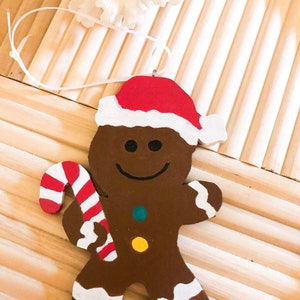 Puede incluir: Un adorno de hombre de jengibre marr&oacute;n con un gorro de Pap&aacute; Noel rojo y blanco. La figura tiene una sonrisa negra, dos peque&ntilde;os botones circulares en color verde azulado y amarillo, y detalles de glaseado blanco en los pies y brazos. Sostiene un bast&oacute;n de caramelo.