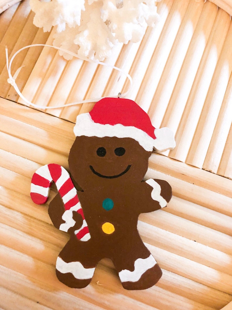 Puede incluir: Un adorno de hombre de jengibre marr&oacute;n con un gorro de Pap&aacute; Noel rojo y blanco. La figura tiene una sonrisa negra, dos peque&ntilde;os botones circulares en color verde azulado y amarillo, y detalles de glaseado blanco en los pies y brazos. Sostiene un bast&oacute;n de caramelo.