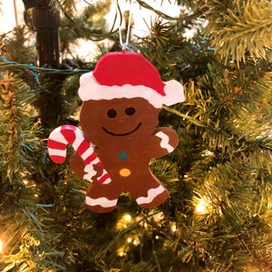 Puede incluir: Un adorno de hombre de jengibre marr&oacute;n con un gorro de Pap&aacute; Noel rojo y blanco, sosteniendo un bast&oacute;n de caramelo a rayas rojas y blancas. La figura presenta una cara sonriente pintada, dos peque&ntilde;os botones de colores y detalles de glaseado blanco.