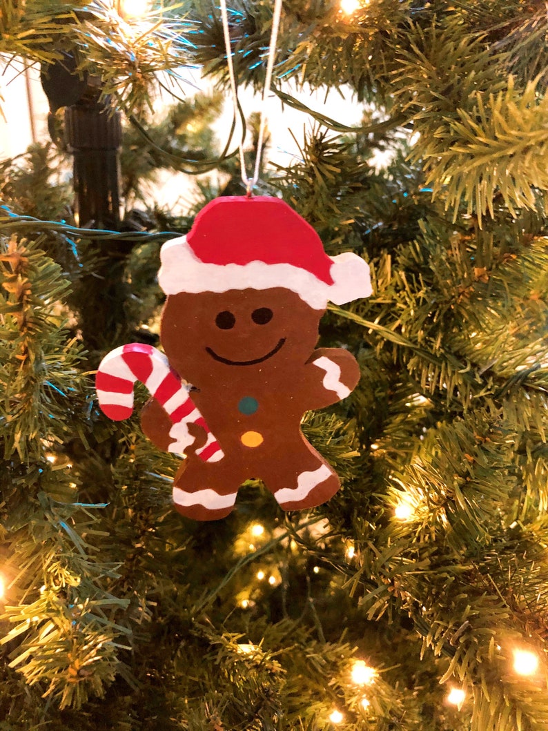 Puede incluir: Un adorno de hombre de jengibre marr&oacute;n con un gorro de Pap&aacute; Noel rojo y blanco, sosteniendo un bast&oacute;n de caramelo a rayas rojas y blancas. La figura presenta una cara sonriente pintada, dos peque&ntilde;os botones de colores y detalles de glaseado blanco.
