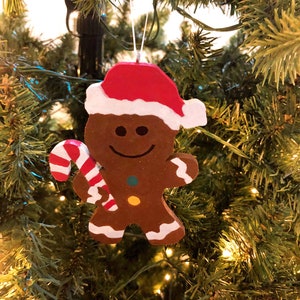 Puede incluir: Un adorno de hombre de jengibre marr&oacute;n con un gorro de Pap&aacute; Noel rojo y blanco, sosteniendo un bast&oacute;n de caramelo a rayas rojas y blancas. La figura presenta una cara sonriente, dos botones coloridos en el torso y detalles de glaseado blanco.