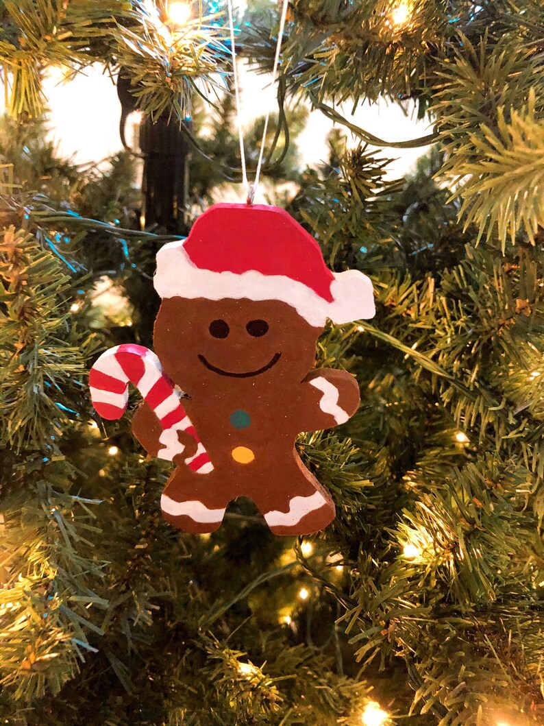 Puede incluir: Un adorno de hombre de jengibre marr&oacute;n con un gorro de Pap&aacute; Noel rojo y blanco, sosteniendo un bast&oacute;n de caramelo a rayas rojas y blancas. La figura presenta una cara sonriente, dos botones coloridos en el torso y detalles de glaseado blanco.
