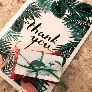 Puede incluir: Un sobre acolchado blanco con un patr&oacute;n de hojas de palma tropicales verdes y flamencos rosas con el texto thank you en letra cursiva negra. Un peque&ntilde;o paquete rojo est&aacute; atado con una cinta verde y una tarjeta de CoastioDesigns.