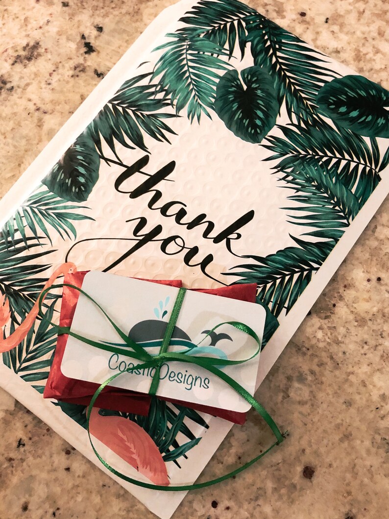 Puede incluir: Un sobre acolchado blanco con un patr&oacute;n de hojas de palma tropicales verdes y flamencos rosas con el texto thank you en letra cursiva negra. Un peque&ntilde;o paquete rojo est&aacute; atado con una cinta verde y una tarjeta de CoastioDesigns.