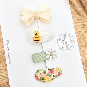 以下が含まれることがあります： 黄色いリボン、蜂の付いた白いクリップ、白い花の付いた緑のクリップ、花柄のクリップの4つのヘアクリップのセットです。クリップは、「Hush-a-bye Bay」というテキストとInstagramのハンドル「@hushabye_bay」が書かれた白いカードに表示されています。