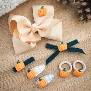 May include: A collection of fall-themed hair accessories, including a large peach-colored bow, green velvet barrettes, and hair ties. Each item is adorned with a small orange pumpkin with a green stem. The accessories are arranged on a wooden surface.