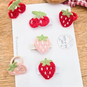 May include: A selection of handmade crochet hair clips shaped like fruits. The clips feature cherries, strawberries, and a small purse. The clips are red, pink, and green, with white detailing. The background includes a white card and wooden surface.