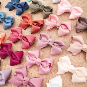 Pode incluir: Uma coleção de laços de cabelo feitos de fita gorgorão canelada em uma variedade de cores, incluindo roxo, azul-petróleo, cinza claro, azul marinho, verde oliva, rosa, laranja, rosa antigo, vermelho escuro, malva, creme e branco.