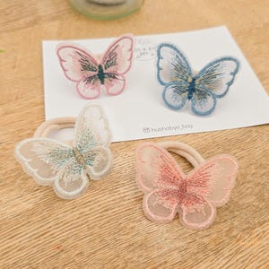 May include: Four butterfly hair ties in pink, blue, and white, with embroidered details. Each butterfly is attached to a beige elastic band. The hair accessories are arranged on a white card and a wooden surface.