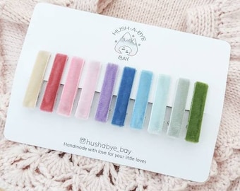 May include: A white card with the text "Hush-A-Bye Bay" and a small mountain illustration. Ten velvet ribbons in various colors are displayed on the card. The text "@hushabye_bay Handmade with love for your little loves" is at the bottom of the card.