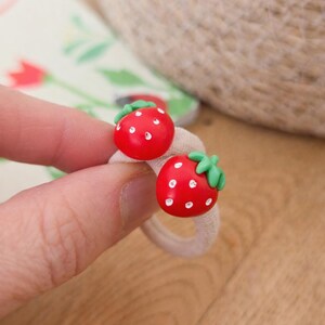 May include: Two red strawberry-shaped hair ties with white dots and green leaves. The hair ties are on a beige elastic band. The background includes a floral pattern and a woven texture.