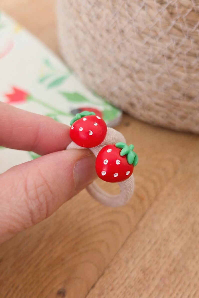 May include: Two red strawberry-shaped hair ties with white dots and green leaves. The hair ties are on a beige elastic band. The background includes a floral pattern and a woven texture.