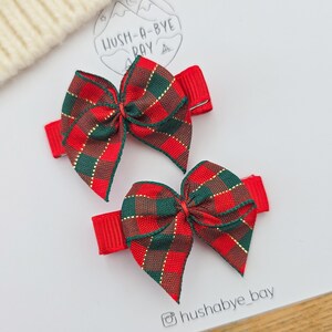 Puede incluir: Dos lazos rojos para el pelo con un estampado a cuadros en verde, marr&oacute;n y dorado. Los lazos est&aacute;n sujetos a pinzas rojas. La imagen tambi&eacute;n incluye el texto "HUSH-A-BYE BAY" y el identificador de Instagram "hushabye_bay".