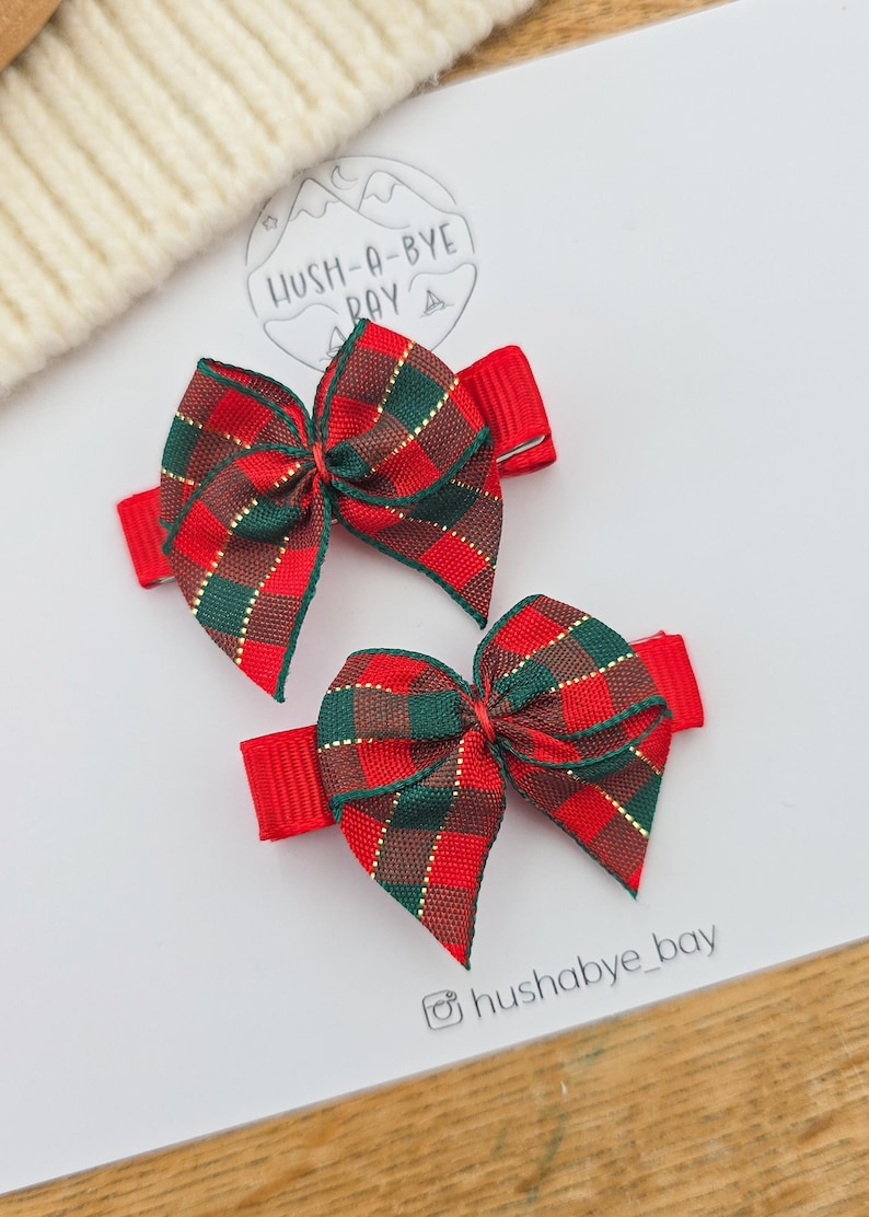 Puede incluir: Dos lazos rojos para el pelo con un estampado a cuadros en verde, marr&oacute;n y dorado. Los lazos est&aacute;n sujetos a pinzas rojas. La imagen tambi&eacute;n incluye el texto "HUSH-A-BYE BAY" y el identificador de Instagram "hushabye_bay".