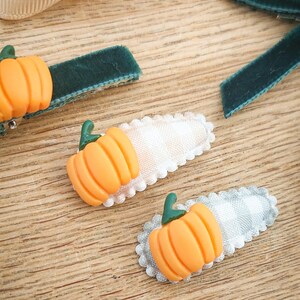 May include: Orange pumpkin hair clips with green stems on a wooden surface. The clips have a white and gray gingham pattern and a scalloped edge. Also, there are green velvet ribbon hair clips.