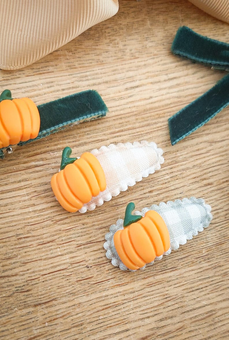 May include: Orange pumpkin hair clips with green stems on a wooden surface. The clips have a white and gray gingham pattern and a scalloped edge. Also, there are green velvet ribbon hair clips.