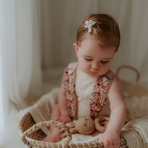May include: A baby girl wearing a floral print romper sits in a woven basket with a crocheted bunny toy. She has a white flower hair clip in her hair.