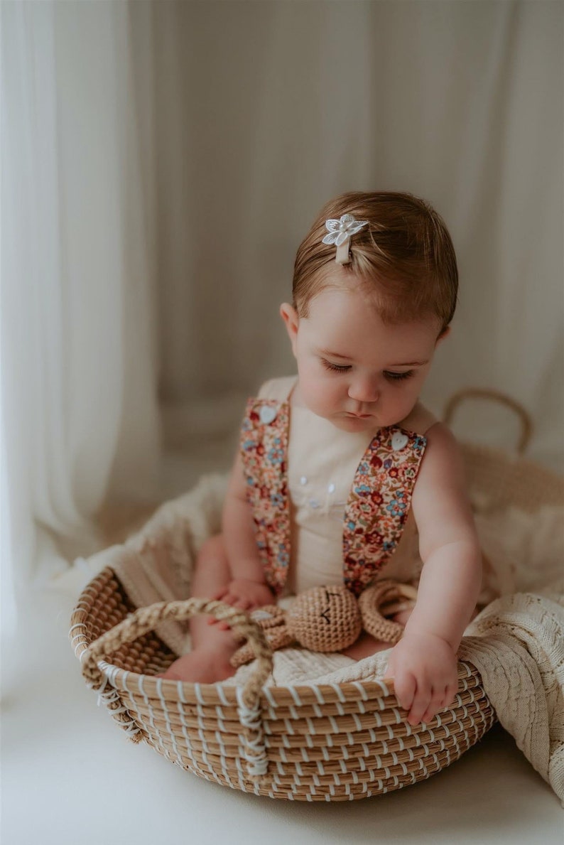 May include: A baby girl wearing a floral print romper sits in a woven basket with a crocheted bunny toy. She has a white flower hair clip in her hair.