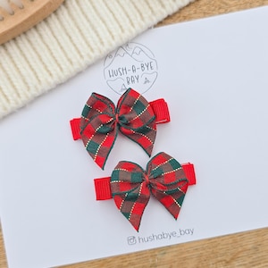 Puede incluir: Dos lazos rojos para el pelo con un estampado de cuadros en rojo, verde y dorado. Los lazos est&aacute;n sujetos a pinzas rojas. La imagen tambi&eacute;n incluye el texto "Hush-A-Bye Bay" y el identificador de Instagram "hushabye_bay".