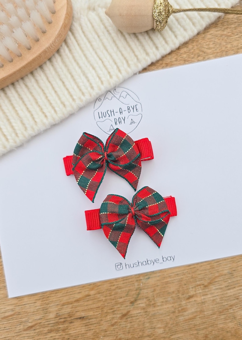 Puede incluir: Dos lazos rojos para el pelo con un estampado de cuadros en rojo, verde y dorado. Los lazos est&aacute;n sujetos a pinzas rojas. La imagen tambi&eacute;n incluye el texto "Hush-A-Bye Bay" y el identificador de Instagram "hushabye_bay".