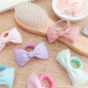 May include: A collection of hair ties featuring small, ribbed fabric bows in pastel colours including pink, lavender, mint green, and light blue. Each bow is attached to a matching elastic band. A wooden hairbrush and a small white dish are visible.