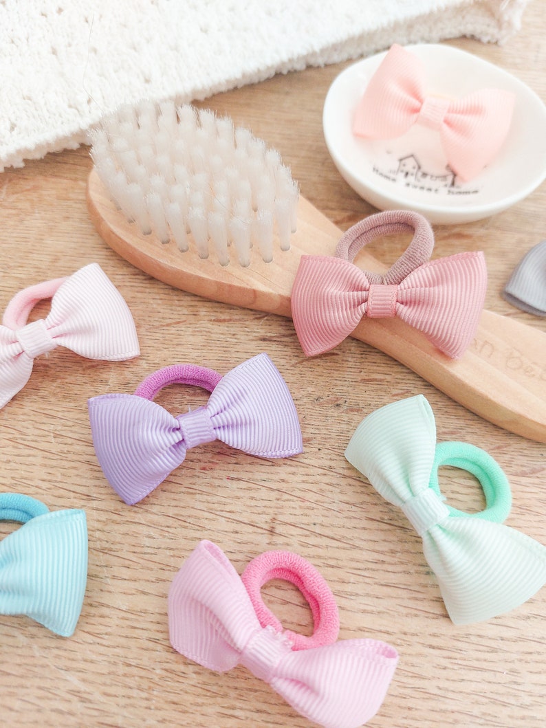 May include: A collection of hair ties featuring small, ribbed fabric bows in pastel colours including pink, lavender, mint green, and light blue. Each bow is attached to a matching elastic band. A wooden hairbrush and a small white dish are visible.
