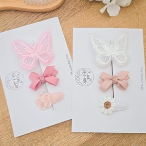 May include: Two sets of baby hair accessories on white cards. One set features a pink butterfly clip, a pink bow clip, and a pink flower clip. The other set includes a white butterfly clip, a pink bow clip, and a white daisy clip. The text "HUSH-A-BYE BAY" is visible.