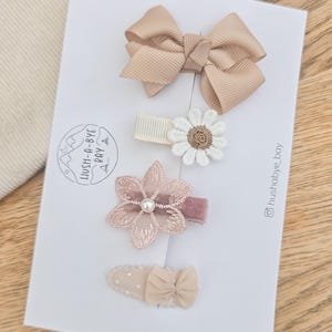 Peut inclure: Un ensemble de quatre barrettes à cheveux sur une carte de présentation blanche marquée Hush-A-Bye Bay. Les accessoires incluent un nœud en ruban gros-grain beige, une marguerite blanche à cœur doré, une fleur en velours mauve et une perle.