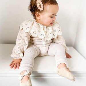 May include: A baby girl wearing a white crocheted lace collar, a beige knitted sweater, and beige striped leggings. She is wearing beige ballet flats with a white bow on the front. She has a pink hair clip in her hair.