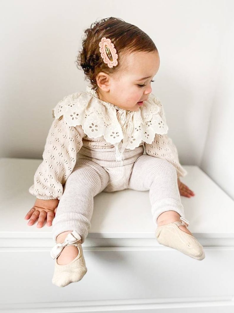 May include: A baby girl wearing a white crocheted lace collar, a beige knitted sweater, and beige striped leggings. She is wearing beige ballet flats with a white bow on the front. She has a pink hair clip in her hair.