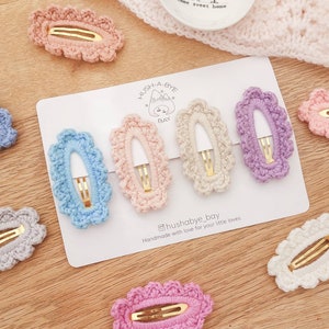 May include: Set of seven crocheted hair clips in various colors, including light blue, pink, purple, beige, and gray. The clips are oval shaped with a scalloped edge and feature a gold metal clasp. The clips are displayed on a white card with the text "Hush-A-Bye Bay" and "Handmade with love for your little loves".