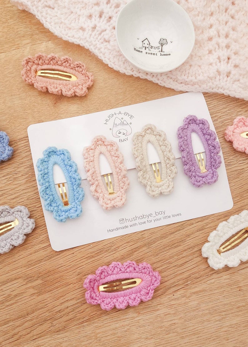 May include: Set of seven crocheted hair clips in various colors, including light blue, pink, purple, beige, and gray. The clips are oval shaped with a scalloped edge and feature a gold metal clasp. The clips are displayed on a white card with the text "Hush-A-Bye Bay" and "Handmade with love for your little loves".