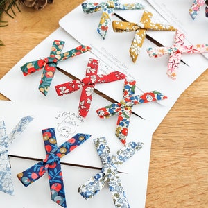 May include: A collection of colorful fabric hair bows in various floral patterns, arranged on white cards. The bows are in shades of red, blue, yellow, and green. The cards have the text "Hush-a-bye Bay" and "Handmade with love for your little loves".