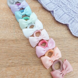 May include: A collection of nine hair ties, each featuring a small, ribbed fabric bow in a different pastel colour, including light blue, lavender, mint green, white, pale pink, soft purple, peach, and dusty rose, arranged in a vertical line.