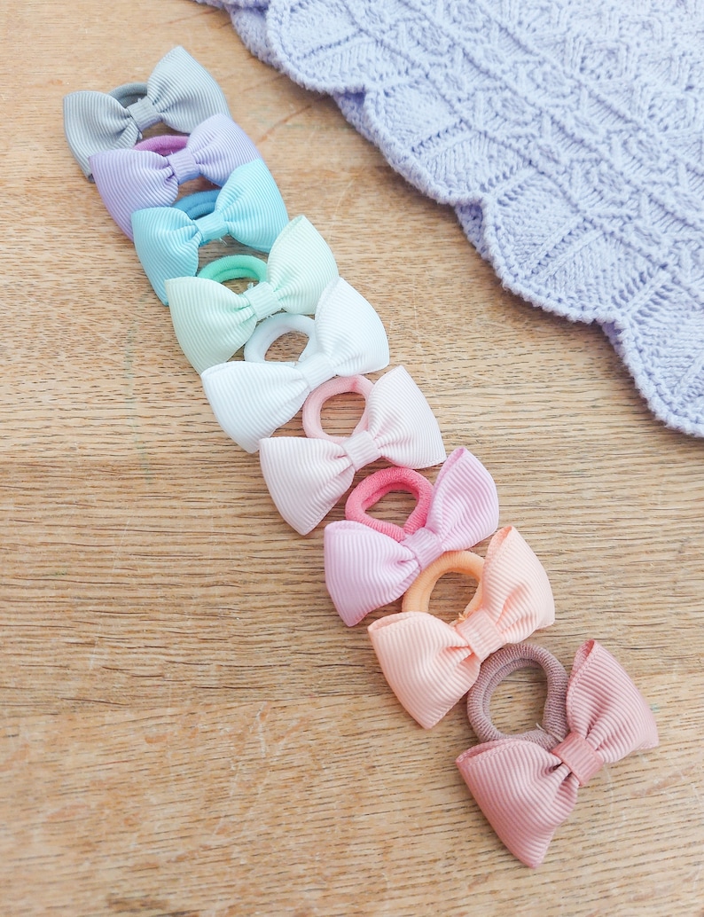 May include: A collection of nine hair ties, each featuring a small, ribbed fabric bow in a different pastel colour, including light blue, lavender, mint green, white, pale pink, soft purple, peach, and dusty rose, arranged in a vertical line.