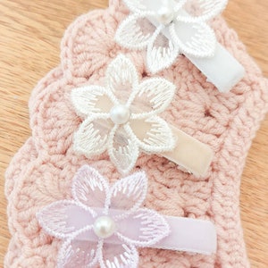 May include: Three white and lavender flower hair clips with pearl centers. The clips are attached to a pink crocheted background.