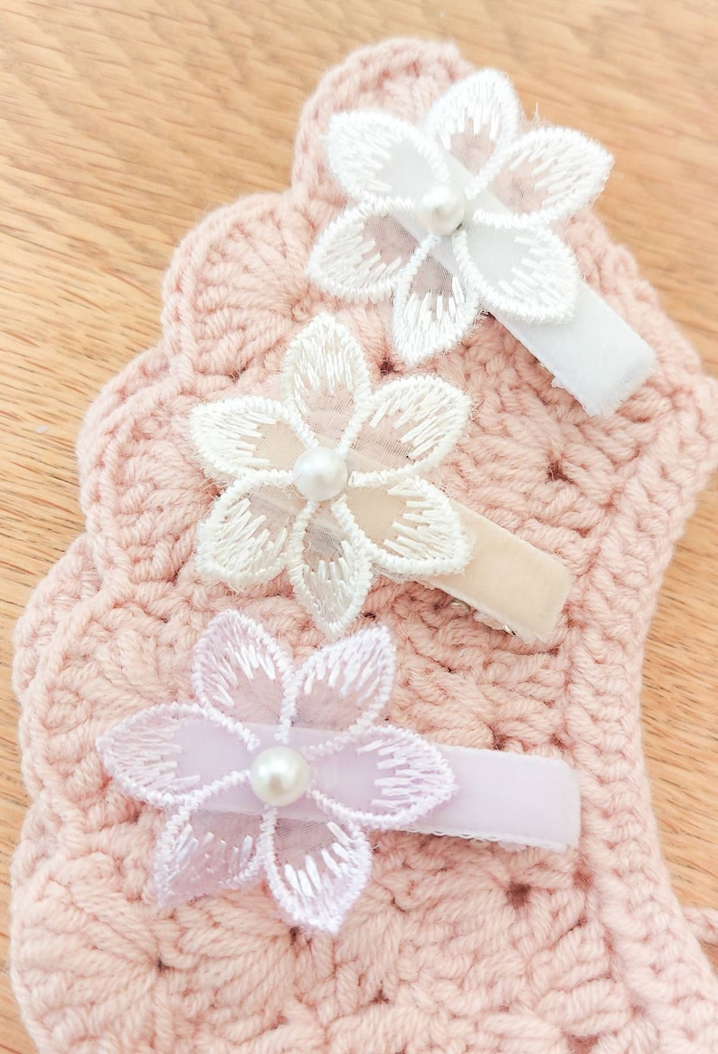 May include: Three white and lavender flower hair clips with pearl centers. The clips are attached to a pink crocheted background.