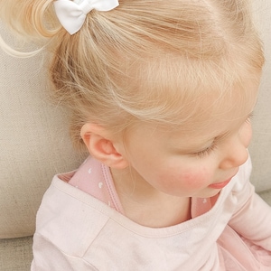 May include: Two small white hair bows with a ribbed texture attached to blonde hair styled in two high pigtails. The hair bows are secured at the base of each pigtail, featuring a simple design with a central knot detail on each white ribbon.