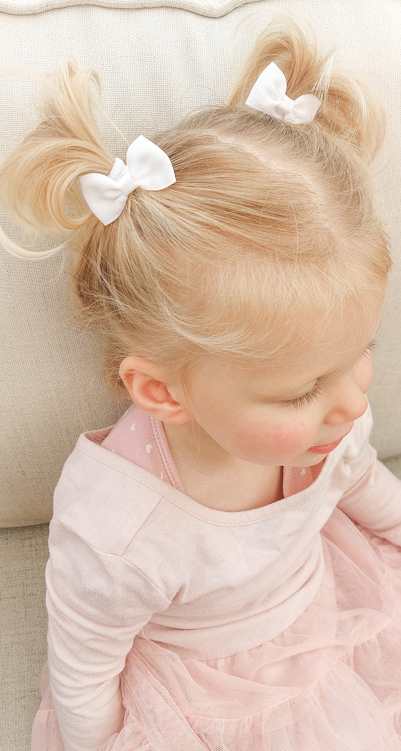 May include: Two small white hair bows with a ribbed texture attached to blonde hair styled in two high pigtails. The hair bows are secured at the base of each pigtail, featuring a simple design with a central knot detail on each white ribbon.