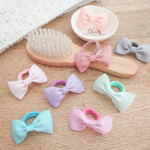 May include: A collection of hair ties featuring small grosgrain ribbon bows in pastel colours including pink, lavender, mint green, light blue, and grey, arranged alongside a wooden hairbrush and a small white ceramic dish with a house illustration.