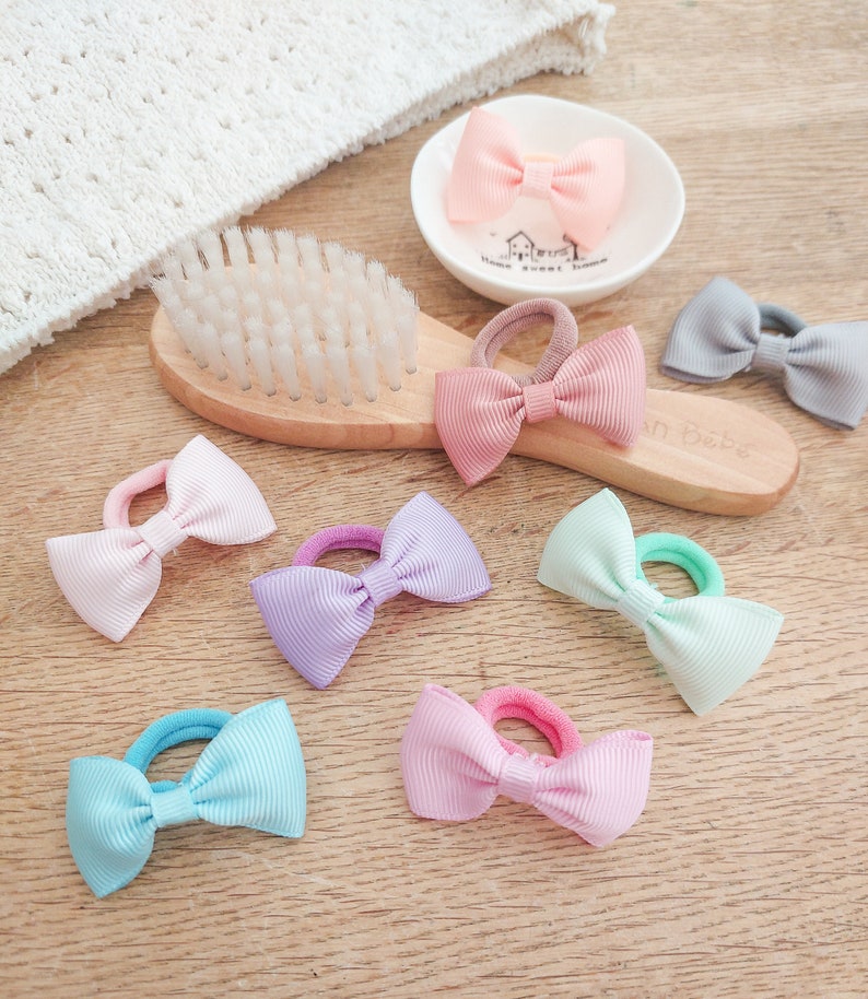 May include: A collection of hair ties featuring small grosgrain ribbon bows in pastel colours including pink, lavender, mint green, light blue, and grey, arranged alongside a wooden hairbrush and a small white ceramic dish with a house illustration.