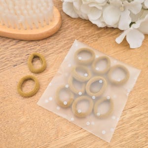 Puede incluir: Un conjunto de gomas para el pelo color beige, algunas sueltas y otras en una bolsa transparente con lunares blancos, est&aacute;n dispuestas sobre una superficie de madera. Tambi&eacute;n se ven un cepillo de pelo de madera y flores blancas.