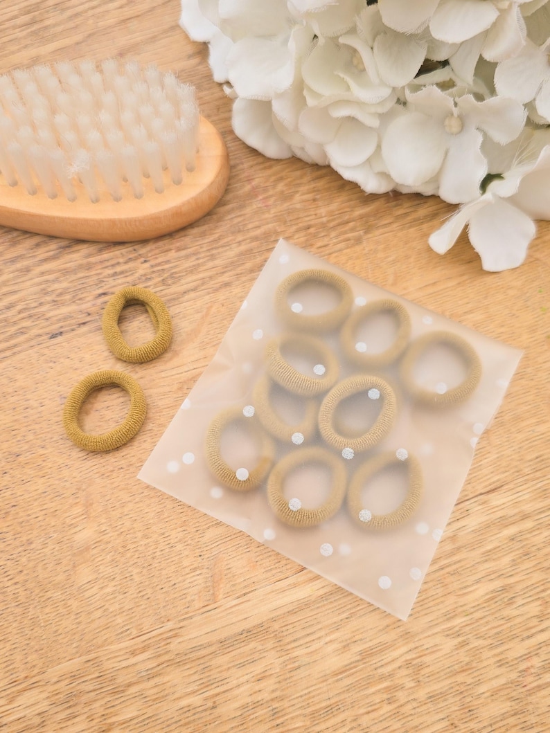 Puede incluir: Un conjunto de gomas para el pelo color beige, algunas sueltas y otras en una bolsa transparente con lunares blancos, est&aacute;n dispuestas sobre una superficie de madera. Tambi&eacute;n se ven un cepillo de pelo de madera y flores blancas.