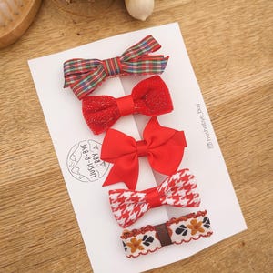 May include: Set of five red and white patterned hair bows. The bows are arranged in a row on a white card with the text "LUSH-A-BYE BABY" and the Instagram handle "@lushbaby_bay".