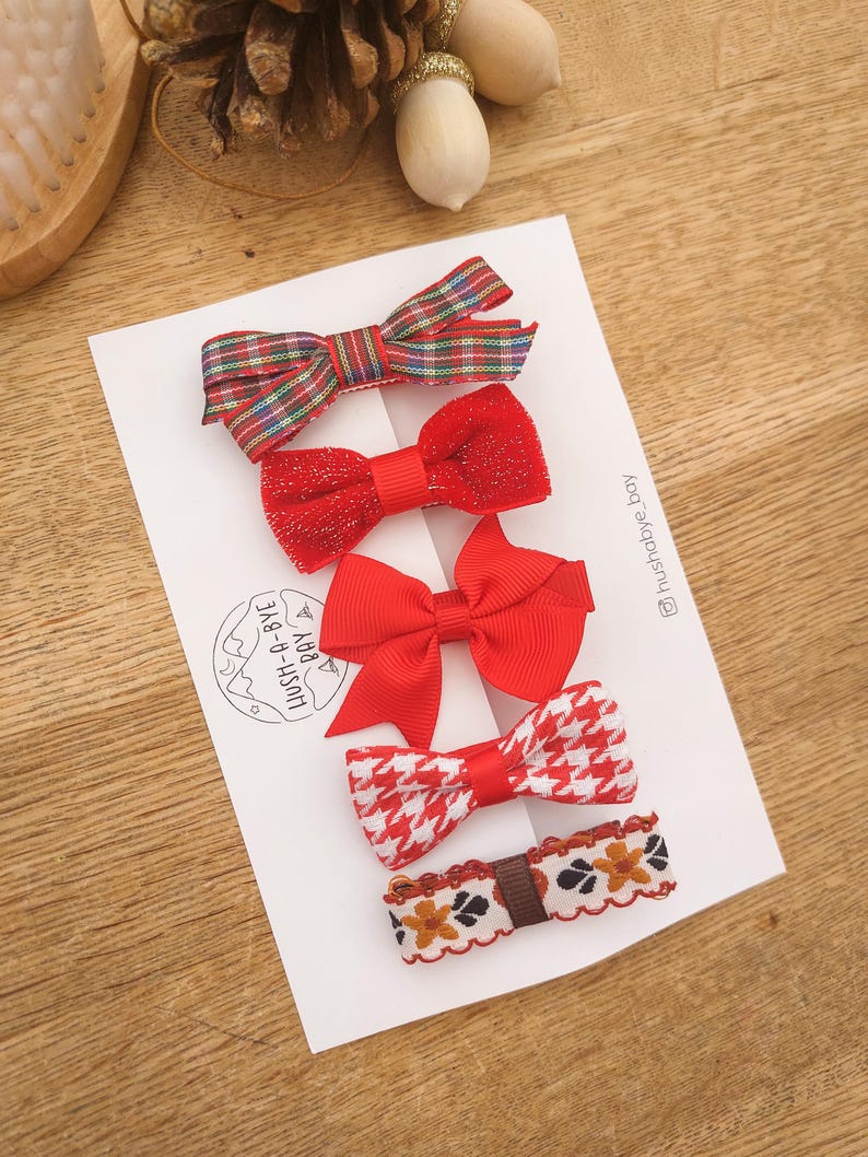 May include: Set of five red and white patterned hair bows. The bows are arranged in a row on a white card with the text "LUSH-A-BYE BABY" and the Instagram handle "@lushbaby_bay".