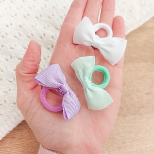 May include: Three hair ties featuring small, ribbed fabric bows in white, mint green, and light purple. Each bow is securely attached to a matching elastic hair band, providing a simple and functional accessory for securing hair in various styles.