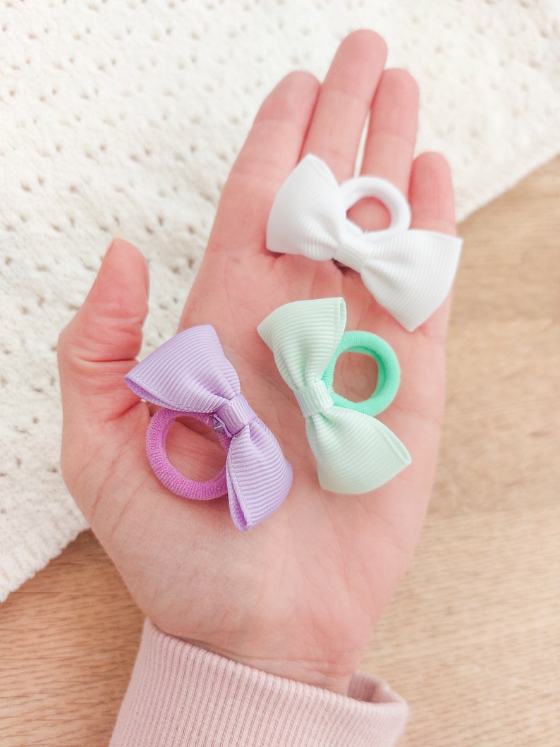 May include: Three hair ties featuring small, ribbed fabric bows in white, mint green, and light purple. Each bow is securely attached to a matching elastic hair band, providing a simple and functional accessory for securing hair in various styles.